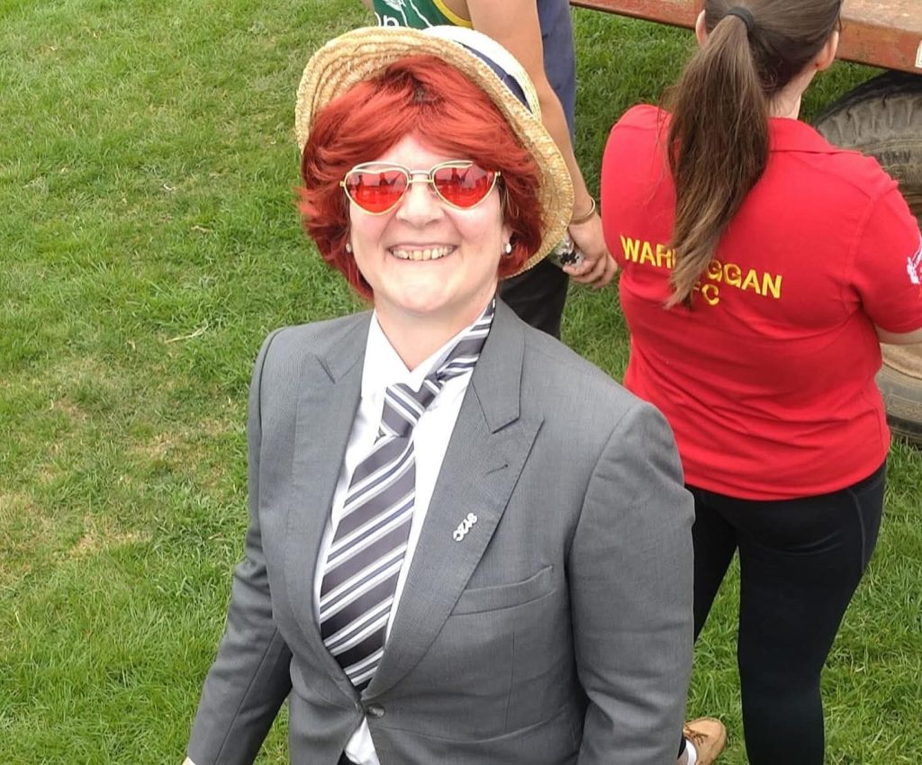 Linda Grant smiling at an outdoor event, wearing a red wig, straw hat and heart-shaped sunglasses
